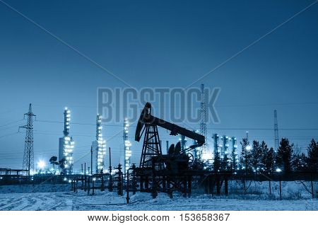 Oil pump and wellhead at the background of refinery by night. Oilfield during winter. Oil and gas industry. Toned.