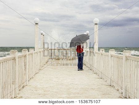 Young girl (brunette) in jeans with a red backpack and a striped umbrella stands on the pier and looking at sea storm. Selective focus.