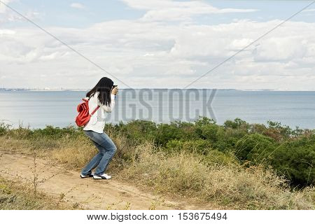 Young girl (brunette) in jeans with a red backpack with a camera taking pictures of the sea. Selective focus.