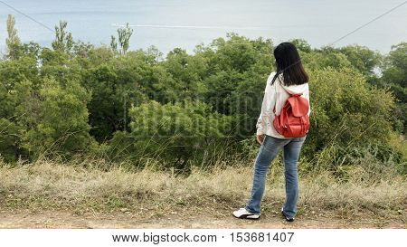 Young girl (brunette) and jeans with a red backpack looking out to the sea. Selective focus.