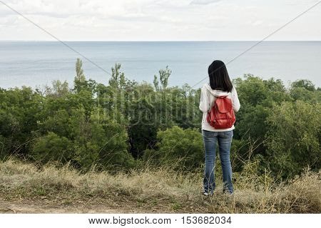 Young girl (brunette) and jeans with a red backpack looking out to the sea. Selective focus.