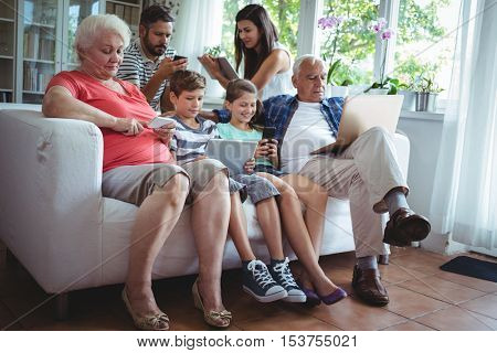 Multi-generation family sitting on sofa and using laptop, mobile phone and digital tablet