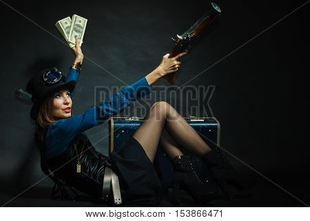 Fashionable subculture weapon concept. Steampunk girl with cash. Young gorgeous lady in victorian fashion lying on floor with banknotes briefcase and blunderbuss.
