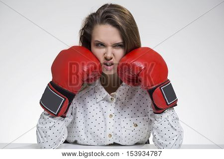 Angry girl in polka shirt is sitting at her table in boxing gloves. Concept of tough business world. Mock up