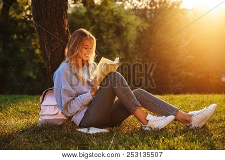Portrait of a delighted young girl sitting on a grass at the park, reading a book, taking notes
