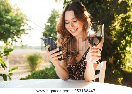 Image of young lady sitting in cafe outdors in park holding glass drinking wine chatting by mobile phone.