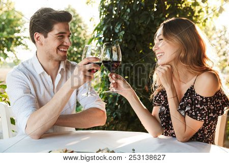 Picture of young loving couple sitting in cafe by dating outdors in park holding glasses of wine drinking.