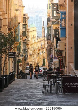 Valletta, Malta - May 2018: Two Girls Walk On Straight Street Of Valletta Between Cafe Chairs