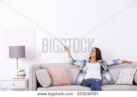 Young Woman Switching On Air Conditioner While Sitting On Sofa Near White Wall