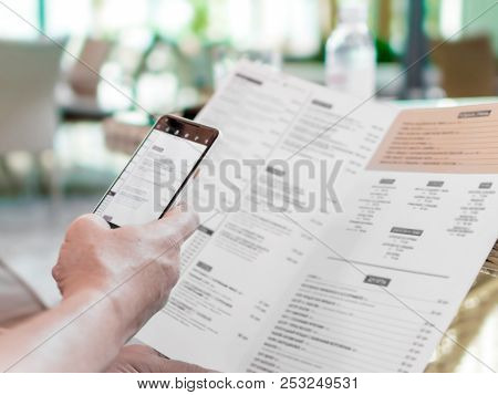 Person's Hands Holding Menu And Mobile Phone In Restaurant. A Foreign Man In Cafe Ordering Food Usin