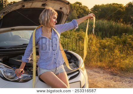 Beautiful Blonde Girl Sits With A Towline On A Hood Of A Broken Car On A Rural Road In The Rays Of T