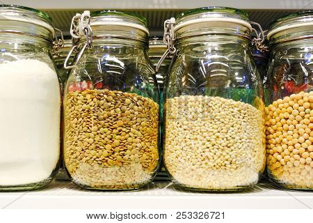 Various Raw Cereals, Grains, Beans And Pasta For Healthy Cooking In Glass Jars On Kitchen Shelf, Whi