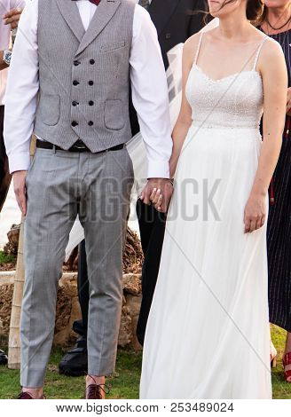 Happy Bride In White With Stones Dress And Bridegroom In Gray Fashionable Suit Holding Hands