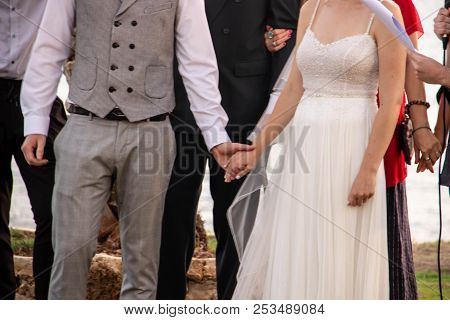 Happy Bride In White With Stones Dress And Bridegroom In Gray Fashionable Suit Holding Hands
