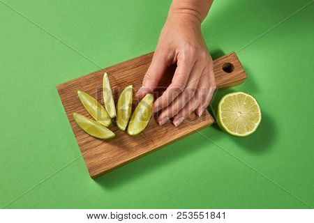 Top view of citrus background. Slices of lime on a board on a green table. A female hand puts slice of ripe fresh green lime on a wooden cutting board with copy space.