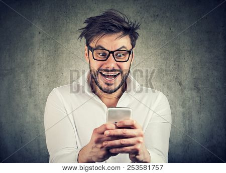 Thrilled And Crazily Looking Young Man In Glasses And Shirt Watching News On Smartphone Standing Aga