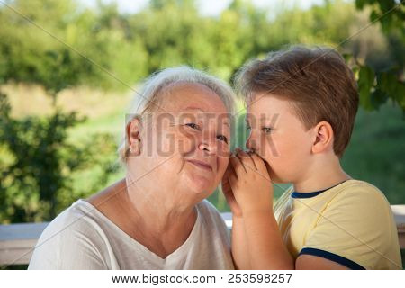 Boy whispering to grandma in summer outdoors garden, grandkid and grandmother lalking.