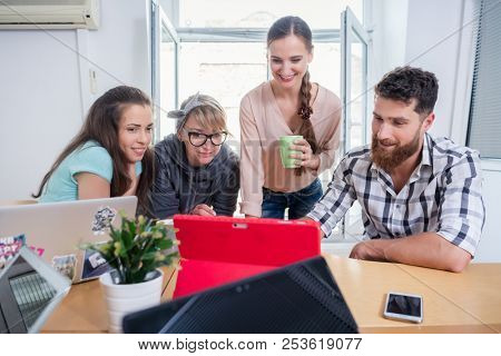 Four co-workers smiling, while watching together a professional business presentation or a funny video on tablet in a modern shared office space for freelancers or young entrepreneurs
