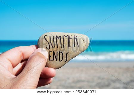 closeup of the hand of a young caucasian man on the beach, in front of the ocean, holding a stone with the text summer ends written in it