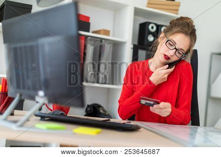 A Young Girl Is Sitting At The Desk In The Office, Holding A Bank Card In Her Hand, Typing On The Ke