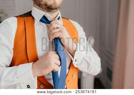 Indoor Shot Of Elegant Male Groom Dressed In Festive Outfit, Ties Tie, Prepares For Special Occasion