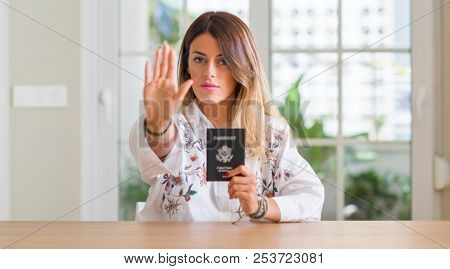 Young woman at home holding a passport of United states with open hand doing stop sign with serious and confident expression, defense gesture