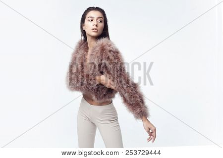 Young Elegant Girl Posing In Fluffy Pink Cape With Artificial Fur And Beige Flare Trousers On Gray B