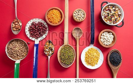 Top View Of Fflat Lay Of Assortment Of Legumes On Red Tabletop Background, In Scoop And Ladles.