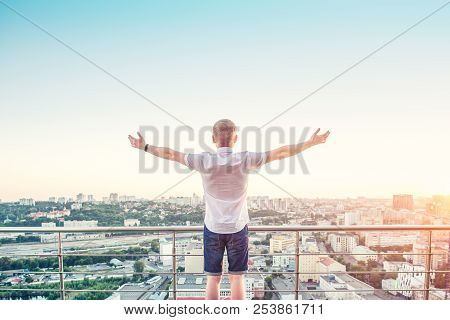 Back View Man On A High Rise Building Balcony Overlooking City With Hands Rised Up To Sky, Feeling A