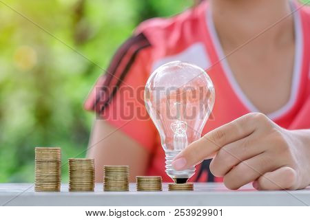 Hand Holding Light Bulb With Coins Stack On Wooden Table In The Morning. Energy And Money Saving , A