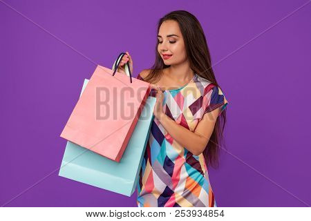 Beautiful Woman Peeks Into A Package, In The Hands Of Multi-colored Shopping Bags On A Purple Backgr
