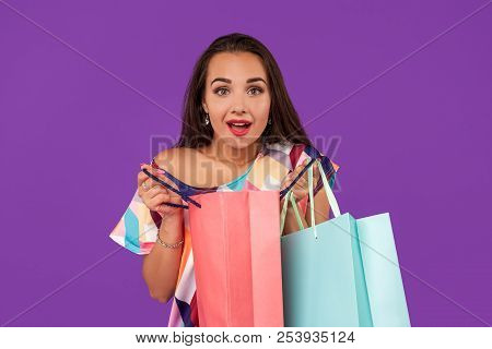 Beautiful Woman Peeks Into A Package, In The Hands Of Multi-colored Shopping Bags On A Purple Backgr