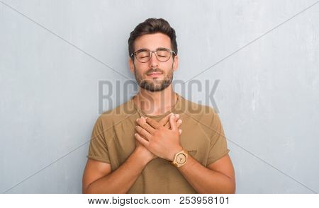 Handsome young man over grey grunge wall wearing glasses smiling with hands on chest with closed eyes and grateful gesture on face. Health concept.