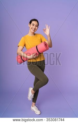 Full length of a cheerful young pretty sportswoman standing isolated over violet background, carrying fitness mat, shosing ok
