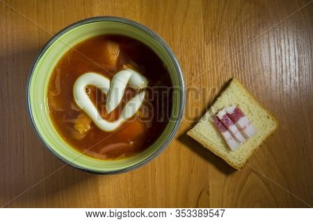 A Bowl Of Red Borsch With Heart-shaped Mayonnaise And A Slice Of White Bread And Slices Of Bacon.