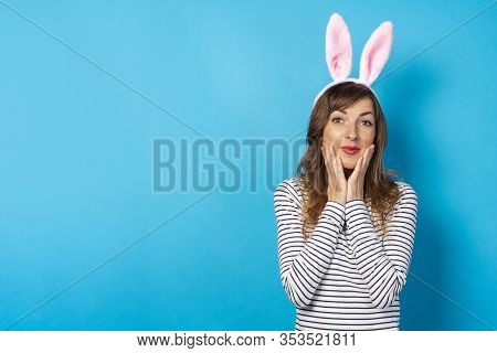 Young Brunette Woman With Rabbit Ears Holds Two Hands In Her Face On A Blue Background With Empty Sp