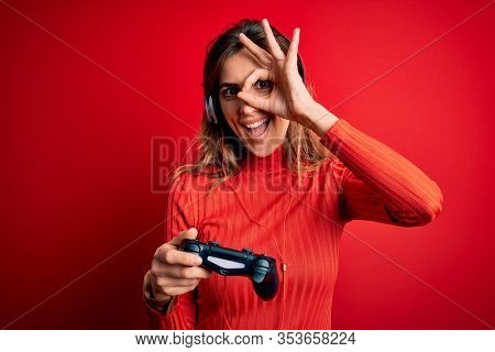 Young beautiful brunette gamer woman playing video game using joystic and headphones with happy face smiling doing ok sign with hand on eye looking through fingers
