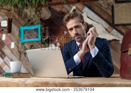young businessman in suit rubbing hands, holding elbows on desk and sitting in a coffeshop