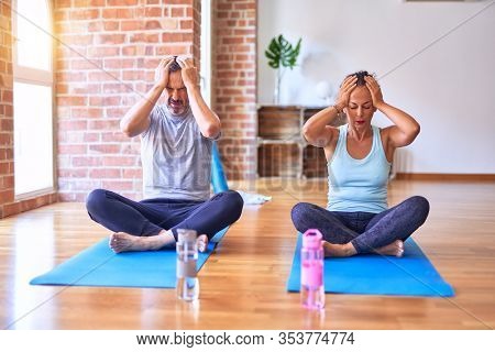 Middle age sporty couple sitting on mat doing stretching yoga exercise at gym suffering from headache desperate and stressed because pain and migraine. Hands on head.