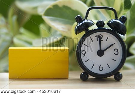 Black Alarm Clock And Blank Yellow Wooden Block Isolated On Blurred Nature Background. The Clock Set