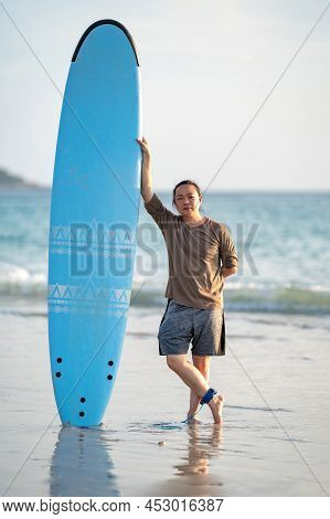 Smart Asian Man Long Hair Touches And Poses With Standed Surfboard On The Beach In Twilight Time