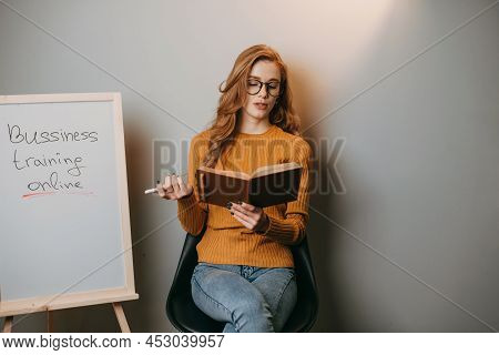 Woman Sitting On Chair Next To A White Board On Which She Writes Business Training Online, Holding I
