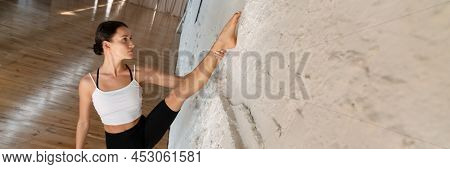 Young brunette white woman in sportswear leaning on a wall stetching leg in studio