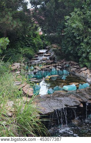 Beautiful Fountain With Rocks And Waterfalls. Fountain In The Park Near Big Green Trees