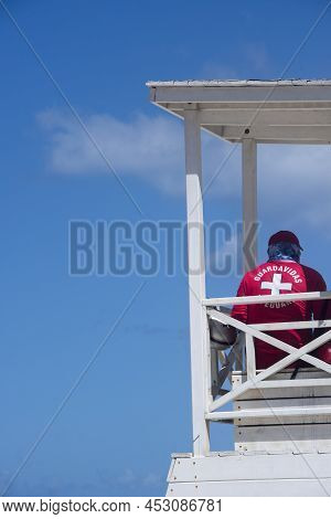 A Lifeguard In A Red Uniform With White Crosses, Watches The Beach From His Tower Against The Blue S