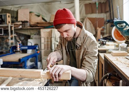 Bearded Caucasian Carpenter Wearing Red Knit Cap Cutting Mortise In Wood Plank Using Chisel
