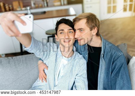Loving Happy Couple Of Homosexual Guys Spend Time Together At Home On The Couch, Take A Selfie On Th