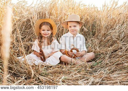 Happy Child Boy And Girl In Straw Hats Sitting In Wheat Field, Eating Bread On Summer. Cottage Core