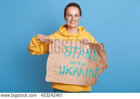 Smiling Delighted Woman In Yellow Hoodie With Ukrainian Flag On Cheeks, Holding Pointing Placard Wit