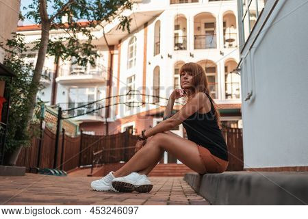 Young Woman With Tattooes Is Sitting On The Sidewalk. Side View. The Concept Of Social Isolation And